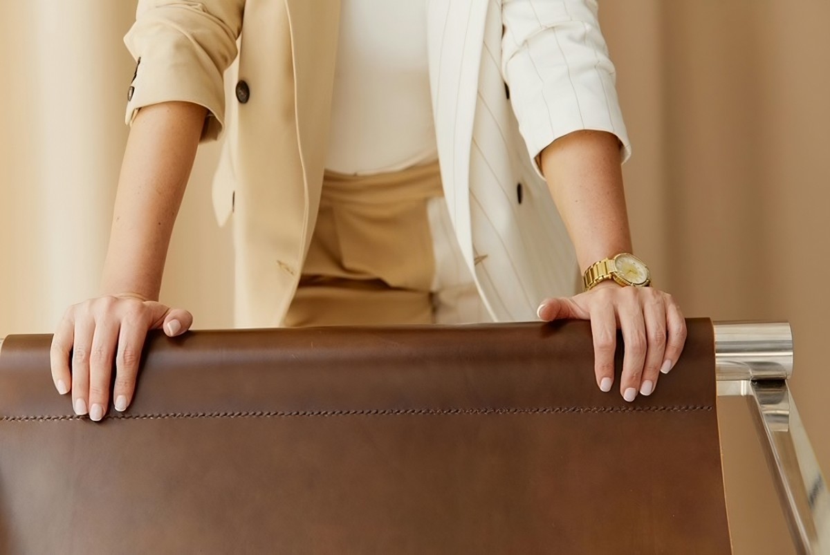 Gros plan sur une femme en tailleur beige et blanc, posant les mains sur un dossier de chaise en cuir, portant une montre dorée élégante à son poignet. Cette image illustre le raffinement et l’importance de choisir une montre comme accessoire de mode chic.